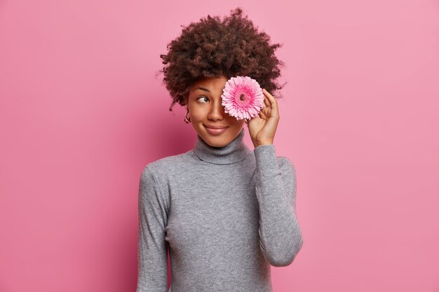 素敵なロマンチックな若い女性の屋内ショットは、目の上に花を保持し、穏やかな笑顔を持って、カジュアルな灰色のタートルネックに身を包んだ