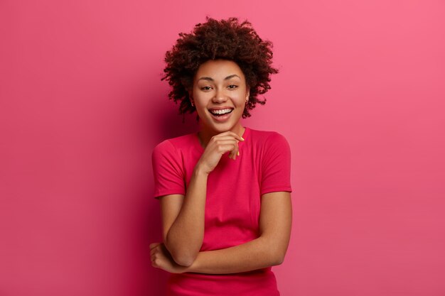 Indoor shot of lovely woman giggles happily, looks straightly , keeps hand under chin, listens hilarious joke, dressed in casual wear, poses against pink wall. Emotions concept