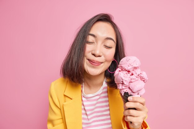 Indoor shot of lovely woman keeps eyes closed from pleasure while eating tasty ice cream being in good mood wears stylish clothing. Beautiful eastern female model poses with delicious frozen snack