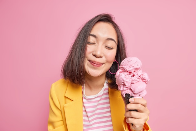 Снимок в помещении: милая женщина держит глаза закрытыми от удовольствия, когда ест вкусное мороженое в хорошем настроении в стильной одежде. Красивая восточная модель позирует с вкусной замороженной закуской