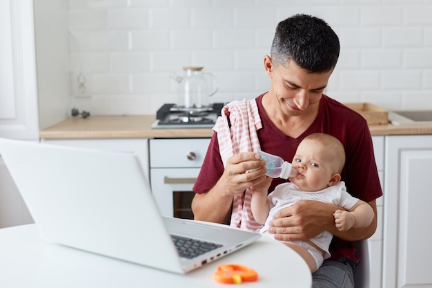 Крытый снимок человека в темно-бордовой повседневной футболке с детской бутылочкой, маленькой дочери или сына, пьющего воду руками отца, семьи, позирующей за столом на кухне.