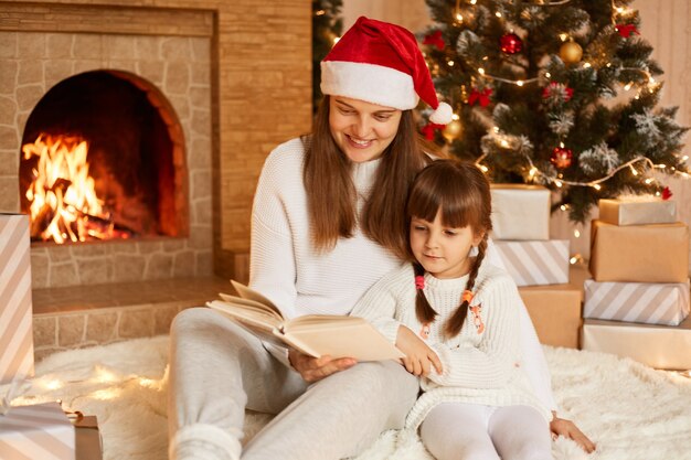 Indoor shot of mother and daughter reading fairy tails in new year eve, woman wearing white sweater and Santa Claus hat readsbook her charming kid, posing in festive room on floor.