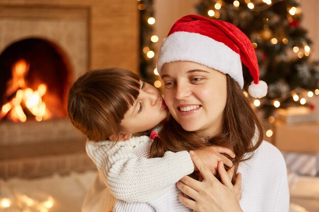 Indoor shot of mother and her little daughter hugging each other, having good mood, little cute girl kissing her mommy, merry Christmas happy new year.