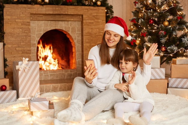 Indoor shot of mother and little daughter having video call or broadcasting livestream, waving hands to smart phone camera, posing near fireplace and Christmas tree.