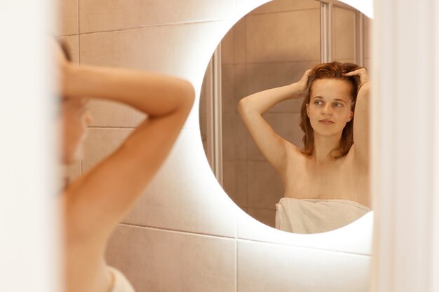 Indoor shot of natural beautiful young adult woman looking at herself reflection in mirror, standing with raised head, doing beauty procedures after taking shower in morning.