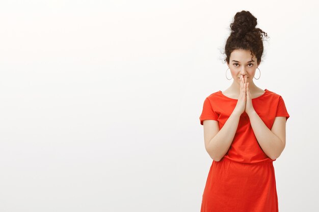 Indoor shot of nervous intense attractive female in red dress with curly hair in bun hairstyle, holding hands in pray over mouth