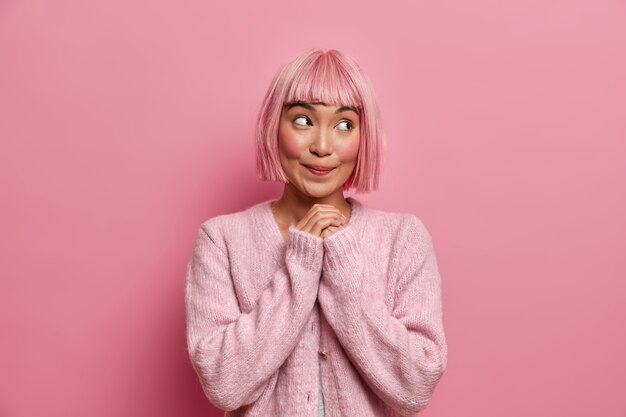 Бесплатное фото Снимок в помещении: красивая мечтательная молодая женщина с короткими розовыми волосами смотрит в сторону со счастливым довольным выражением лица, держит руки вместе, носит джемпер