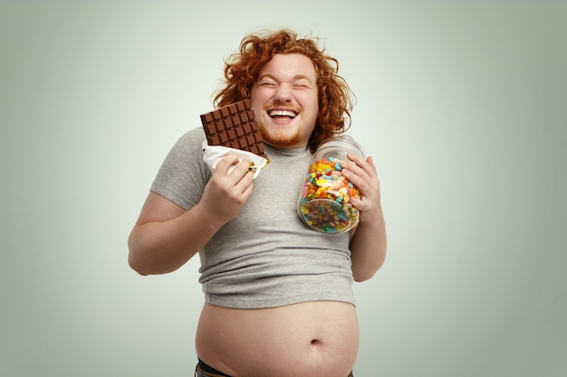 Бесплатное фото Снимок в помещении: толстый рыжий мужчина держит в одной руке стакан конфет, а в другой - плитку шоколада