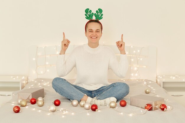 Крытый снимок счастливой позитивной женщины в белом пуловере, джинсах и оленьем обруче на рождественской вечеринке, сидящей на кровати в спальне и указывающей вверх указательными пальцами.