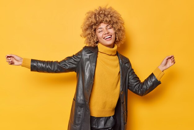 Бесплатное фото Снимок счастливой женщины в помещении трясет руками, танцует беззаботно, держит глаза закрытыми, улыбается зубами, носит свитер и кожаное черное пальто, изолированное на желтом фоне.