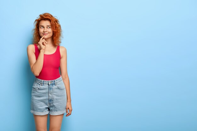 Бесплатное фото Снимок в помещении: счастливая женщина с рыжими волосами с задумчивым выражением лица, одетая в повседневный жилет и джинсовые шорты, представляет что-то приятное