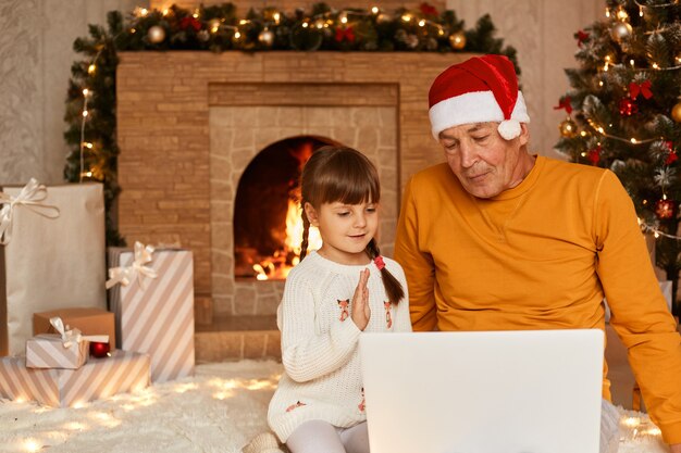オレンジ色​の​セーター​と​サンタクロース​の​帽子​を​かぶった​成熟した​男性​と​、​ラップ​トップ​の​前​に​座って​ビデオ​通話​を​し​、​クリスマスツリー​と​リビング​ルーム​の​暖炉​の​近く​で​ポーズ​を​とっている​小さな​かわいい​黒髪​の​女の子​の​屋内​ショット​。