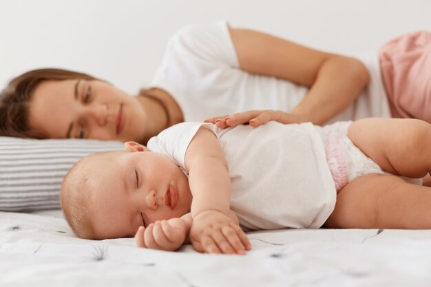 웃고 있는 여성이 잠자는 딸과 함께 누워 사랑스러운 아기와 함께 쉬고 있는 실내 사진, 흰색 캐주얼 티셔츠를 입은 여성이 긍정적인 감정으로 어린 소녀를 바라보고 있습니다.