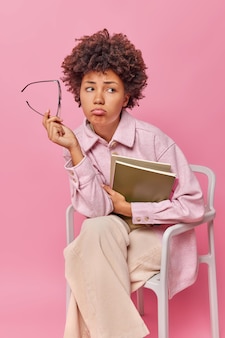 공부하는 데 지친 불행한 여학생의 실내 사진은 안경을 벗고 교과서를 들고 메모장은 의자에 편안하게 앉아 분홍색 벽에 격리된 평상복을 입고 있습니다.