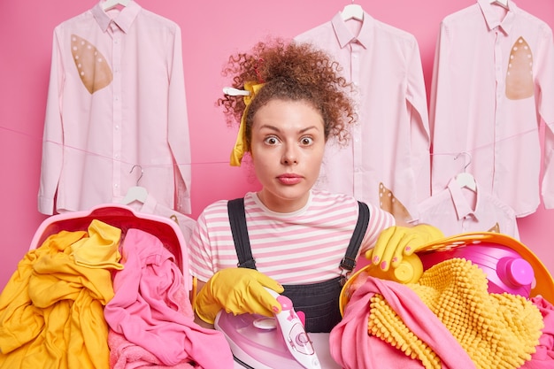 Бесплатное фото Снимок в помещении молодой девушки с кудрявыми волосами, окруженной корзиной с утюгом для стирки, вымытой линией, удивил, глядя на гладильную доску, позирующую на розовой стене, занятую поглаживанием вещей