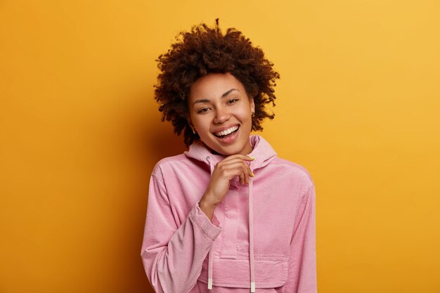 Indoor shot of optimistic curly haired pretty woman keeps hand under chin, looks directly , being in good mood, stands satisfied and delighted, wears casual sweatshirt, poses indoor