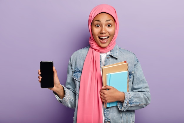 Indoor shot of optimistic dark skinned Muslim woman introduces cool gadget