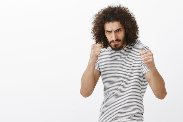 Tiro al coperto di bullo indignato con barba e taglio di capelli afro, in piedi nella posa di boxe con i pugni chiusi, aggrottando la fronte al nemico, essendo pronto a combattere