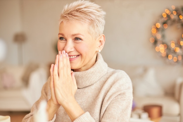 Indoor shot of overjoyed cheerful mature short haired woman in stylish sweater holding hands pressed together at her mouth, smiling broadly, receiving New Year's gift, can't hide her excitement