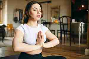 Free photo indoor shot of peaceful young female sitting at home in meditative posture with eyes closed, making namaste gesture, meditating, focusing mind on breathing, training attention and awareness