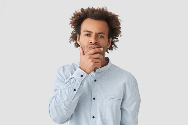 Indoor shot of pensive African American guy holds chin and looks with serious expression, thinks over his problem, wears elegant-shirt, has Afro curly hairstyle, isolated