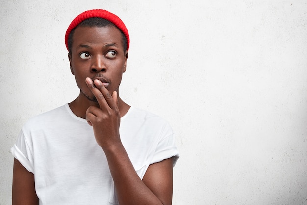 Free photo indoor shot of pensive dark skinned man in trendy clothing, keeps fingers on mouth, thinks about something while looks upwards, stands against white concrete wall