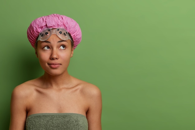 Free photo indoor shot of pensive dark skinned model going to swimming pool, wears water protective hat, rubber goggles and bath towel around body, thinks about something, isolated on green wall.