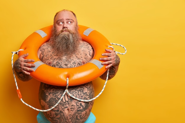 Indoor shot of pensive overweight man looks away, ready for recreation, swimming in sea with lifebuoy, has naked body, isolated on yellow wall, blank space aside. Safety equipment, rescue