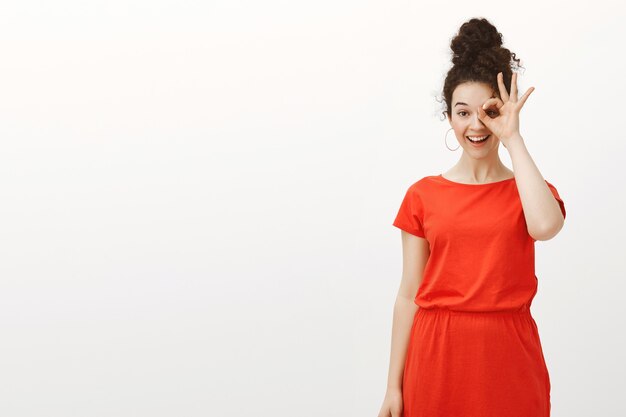 Indoor shot of playful good-looking feminine woman with curly hair in red dress showing okay or approval gesture over eye and smiling broadly