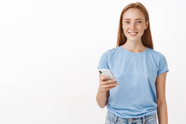 Indoor shot of pleasant happy and friendly redhead woman with freckles in blue casual t-shirt holding smartphone and smiling politely writing down phone number standing over gray wall