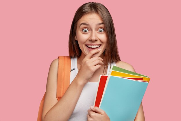 Indoor shot of pleasant looking cheerful woman with toothy smile, keeps hand on chin, looks in amazement, dressed in casual outfit, carries books