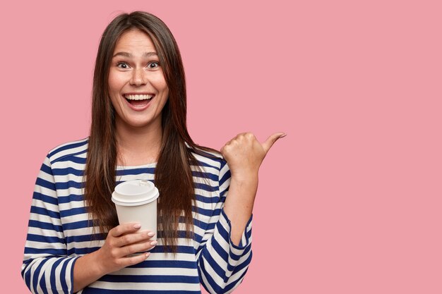 Indoor shot of pleasant looking positive woman with satisfied expression, holds disposable cup of beverage