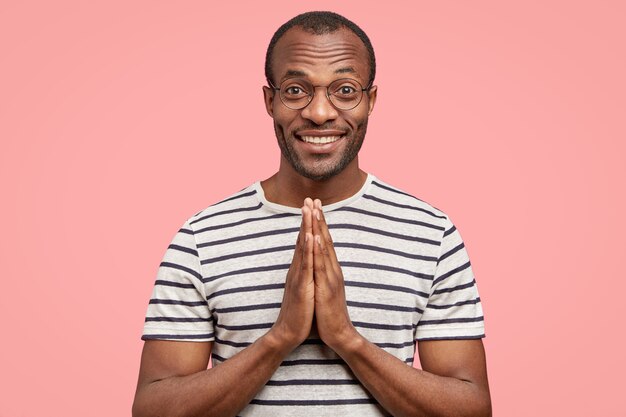 Indoor shot of pleased black dark skinned man keeps both hands in praying gesture