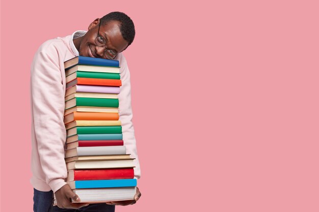 Indoor shot of pleased dark skinned man leans at pile of books, dressed in casual sweater, wears round spectacles