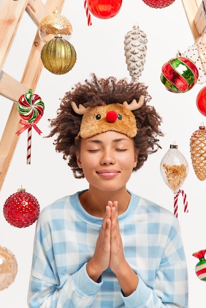 Free photo indoor shot of pleased happy young woman keeps palms in pray gesture makes wish on new year eve closes eyes and smiles gently stands