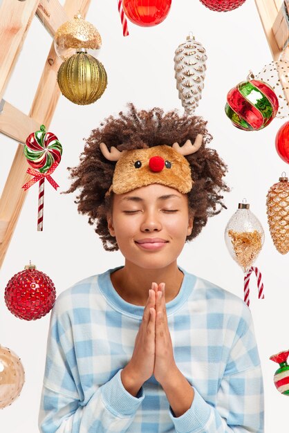 Снимок довольной счастливой молодой женщины в помещении, держащей ладони в жесте молитвы, загадывает желание в канун Нового года, закрывает глаза и нежно улыбается