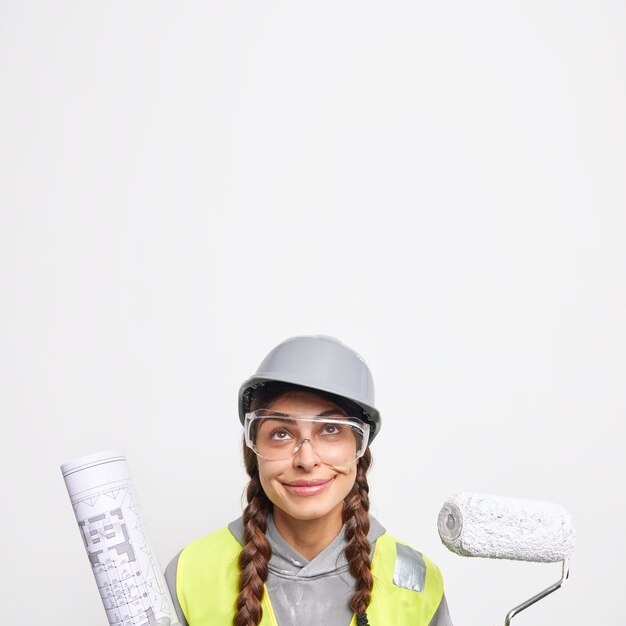 Indoor shot of pleased woman engineer holds painting roller and paper blueprint looks above works on construction site 