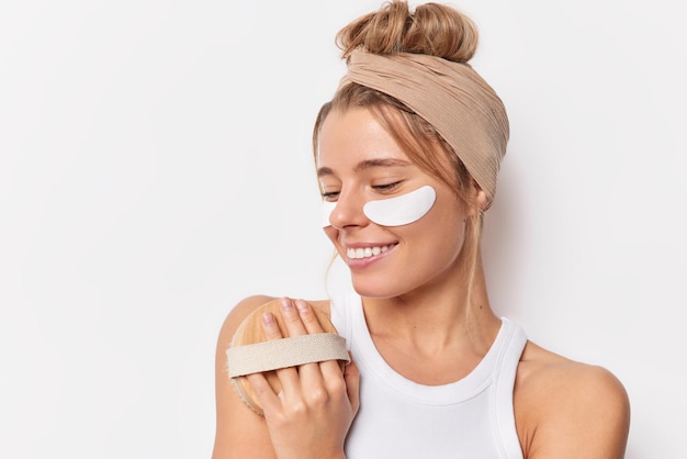 Indoor shot of pleased young cheerful woman massages body wih brush applies beauty patches under eyes undergoes skin care treatments wears headband isolated over white background. Body care concept
