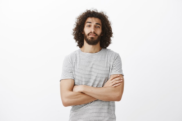 Indoor shot of popular handsome male model with beard and afro hairstyle, holding hands crossed on chest and looking with raised chin