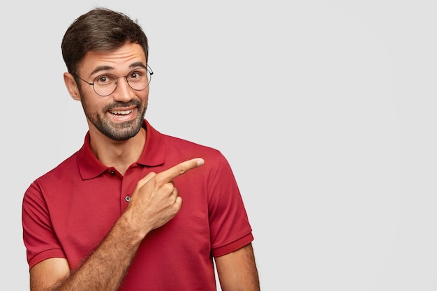 Indoor shot of positive bearded male in casual red t-shirt, points with index finger aside