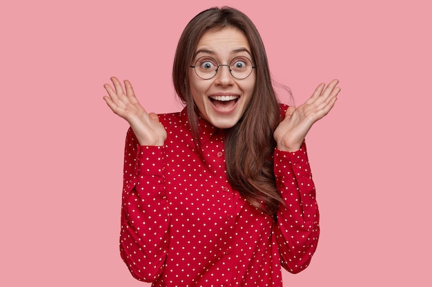 Foto gratuita tiro al coperto di donna bruna positiva in camicia rossa a pois, tiene le mani vicino al viso, si sente felice, ha un'espressione facciale felicissima, modelli su sfondo rosa