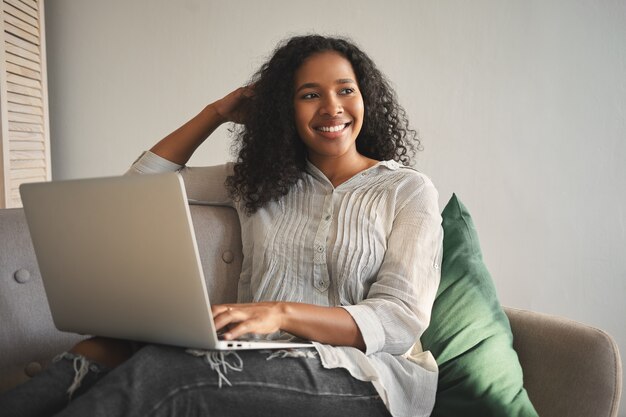 Indoor shot of positive charming young African American woman dressed in stylish clothes relaxing on sofa with portable computer on her lap, shopping online, looking away with cute cheerful smile