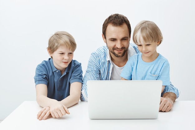 Indoor shot of positive happy blond child with father and brother sitting at table, looking at laptop screen and smiling broadly