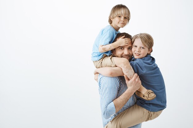 Indoor shot of positive happy family guy holding son with vitiligo on shoulders and cute kid on chest, smiling broadly, feeling joyful while playing with loving children