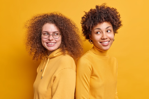 Снимок в помещении: позитивные многонациональные женщины стоят плечом к плечу, улыбаются, красиво одеты, небрежно, вьющиеся волосы в хорошем настроении, изолированы за желтой стеной. Разнообразные счастливые друзья
