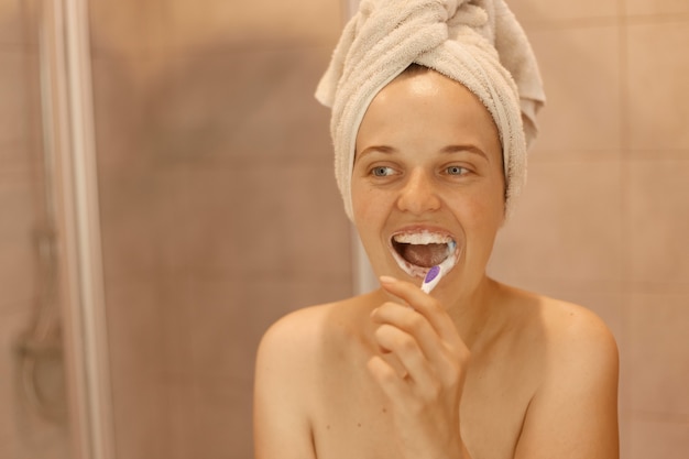 ポジティブで楽観的な美しい女性が歯を磨き、自宅のバスルームでシャワーを浴びた後、白いタオルを頭にかぶって立っている衛生手順を撮影した屋内ショット。