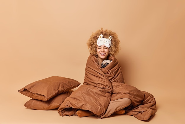 Снимок в помещении позитивной женщины с вьющимися густыми волосами, завернутой в одеяло, держит свою любимую собаку-мопса, широко улыбающуюся, позирует с домашним питомцем в спальне, изолированной на коричневом фоне