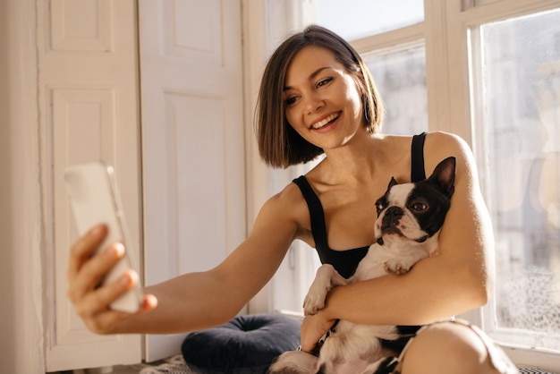 ペットと飼い主のコンセプトの間の愛スマートフォンで自分撮りを保持しているポジティブな若い白人女性の屋内ショット