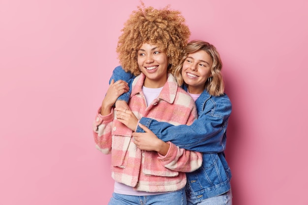 Indoor shot of positive young female friend embrace and have good relationships dressed in fashionable clothes look gladfully into distance isolated over pink background Friendship concept