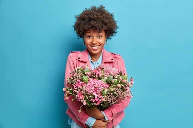 ポジティブな若い女性の屋内ショットは、青い壁に隔離されたピンクのジャケットに心地よく身を包んだ花の笑顔の大きな束を抱きしめます